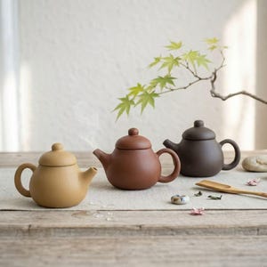 以下が含まれることがあります： 木製の表面に、ベージュ、茶色、濃い茶色の3つのセラミックティーポットが配置されています。各ティーポットは丸みを帯びたボディ、湾曲したハンドル、蓋が付いています。ベージュのティーポットの注ぎ口から蒸気が出ています。木のスプーンと装飾要素も存在します。