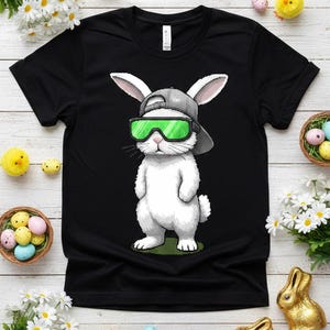 Könnte beinhalten: Schwarzes T-Shirt mit einem Cartoon-Hasen mit grauer Kappe und grüner Sonnenbrille. Der Hase steht auf grünem Gras. Ostermotiv.