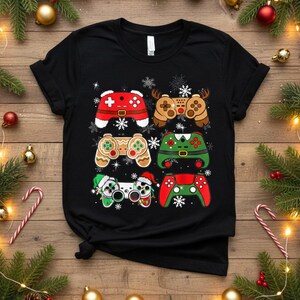 May include: Black t-shirt featuring six festive video game controller designs. The controllers are decorated with holiday themes, including reindeer antlers, Santa hats, and gingerbread men. Snowflakes are scattered around the controllers.