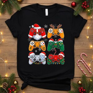 May include: Black t-shirt featuring six colorful video game controllers with Christmas-themed designs. Each controller has a Santa hat, snowflakes, or festive lights. The controllers are in red, orange, green, and gray.