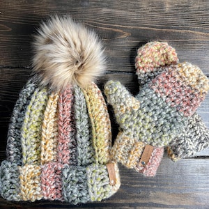 May include: A crochet hat and mittens set in a mix of colours including grey, yellow, and pink. The hat has a ribbed design and a faux fur pom-pom. The mittens and hat have leather tags.