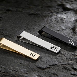 Könnte beinhalten: Drei Metall-Krawattenklammern in Gold, Silber und Schwarz. Jede Klammer hat eingravierte Initialen: HL, MR und CK. Die Krawattenklammern werden auf einer dunklen, strukturierten Oberfläche präsentiert.