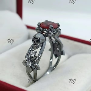 May include: A dark gray ring featuring a skull and floral design. The ring has a large, oval-cut, red gemstone at the top. The band is adorned with a skull, flowers, and small, clear gemstones. The ring is set in a red jewelry box.
