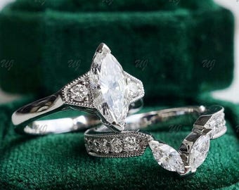 May include: Two silver engagement rings with clear marquise-cut and round-cut stones. The larger ring features a prominent marquise-cut center stone, while the smaller ring has a curved band with smaller stones. Set against a dark green velvet background.