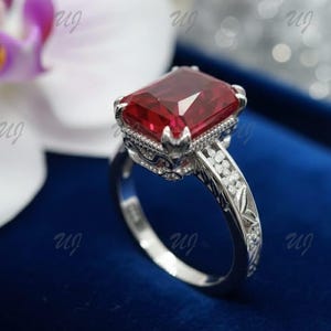 May include: A silver ring featuring a large, rectangular, deep red gemstone. The ring has an ornate, detailed setting with small accent stones and floral engravings on the band. The ring is displayed on a blue surface.