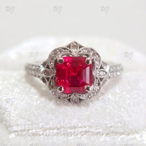 May include: A silver ring featuring a vibrant, square-cut, red gemstone. The central stone is encircled by a halo of smaller, clear gems and delicate filigree. The ring's band is also embellished with tiny, clear stones.