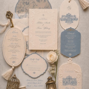 May include: A wedding stationery set featuring cream and blue cards with floral illustrations, ornate borders, and text including Melisa Arada and Bora Yilmaz. Includes a brass key, a small birdcage, a lantern, two wax seals, and a cream-colored rose.