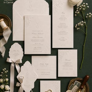 Może przedstawiać: Elegancki zestaw zaproszeń ślubnych w kolorze kości słoniowej z wytłoczonymi detalami kwiatowymi. Zawiera zaproszenie, menu, RSVP i karty ze szczegółami, a także kopertę, ułożone na ciemnozielonej powierzchni. Złoty pierścionek i pieczęć woskowa.