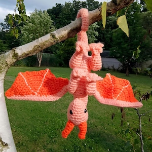 Puede incluir: Un pterodáctilo de ganchillo, rosa y naranja, colgado de una rama de árbol. Las alas son de color naranja brillante con bordes rosas. El juguete tiene pequeños ojos negros y un pico y patas naranjas. El fondo muestra follaje verde.
