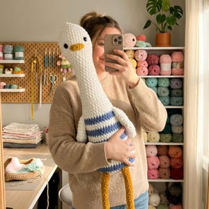 May include: A large, handmade crochet goose doll. The goose has a white body, blue and white striped shirt, yellow feet, and a yellow beak. The doll is being held in front of a mirror, with a craft room in the background.