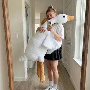 Puede incluir: Un gran peluche de ganso blanco hecho a mano con ganchillo, con pico y patas amarillas. El ganso tiene las alas extendidas y está siendo sujetado. El peluche es un artículo suave y texturizado, perfecto para una habitación infantil o como pieza decorativa.