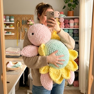 Puede incluir: Un gran peluche de girasol hecho a mano con ganchillo en colores pastel. El peluche presenta una cabeza rosa, un centro azul claro y pétalos amarillos. El juguete se sostiene frente a un espejo.