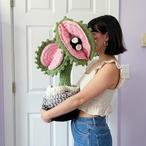 Könnte beinhalten: Eine große, handgefertigte Häkelpflanze mit grünem Stiel und Blättern sowie rosa und weißen Blütenblättern. Die Pflanze steht in einem schwarzen, weißen und grauen Topf. Die Pflanze ist etwa 61 cm hoch.