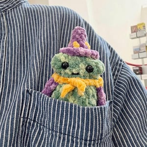 Könnte beinhalten: Eine handgefertigte grüne und lila Häkelfroschfigur mit Hexenhut und Schal, die in einer blau-weiß gestreiften Hemdtasche sitzt. Der Frosch hat schwarze Knopfaugen und einen gelben Schal.