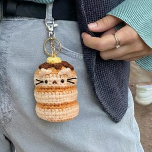 Puede incluir: Un llavero de ganchillo hecho a mano con forma de pila de panqueques con diseño de cara de gato. El llavero es en tonos beige, marrón y amarillo, con un llavero dorado. El llavero cuelga de un pantalón gris.