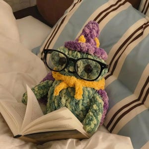 Puede incluir: Una criatura de ganchillo hecha a mano con gafas negras grandes, un sombrero morado y amarillo y una bufanda amarilla, leyendo un libro. La criatura es verde y azul, y está en una cama con almohadas a rayas.