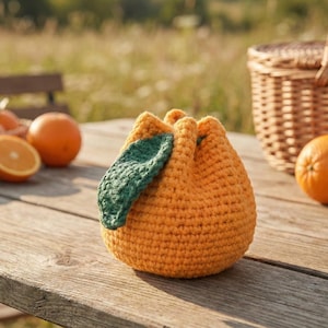 May include: A crocheted orange-colored pouch with a green leaf accent, resembling an orange fruit. The pouch is sitting on a wooden surface with oranges and a wicker basket in the background. The pouch is a unique handmade item.