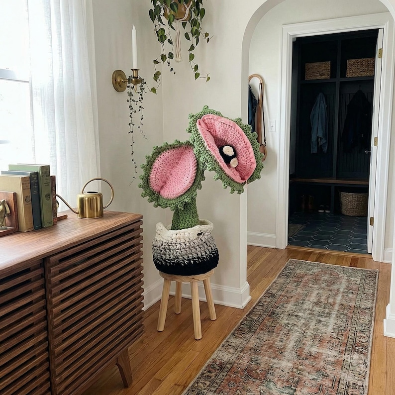 May include: A whimsical, handmade crochet plant with pink and green flower-like structures, set in a black, white, and gray pot. The plant sits on a small wooden stool. The scene is set in a well-lit hallway with a wooden cabinet and a patterned rug.