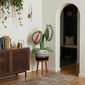 May include: A large, handmade crochet plant with green leaves and pink and green flower heads. The plant sits in a black, white, and gray pot on a small wooden stool. The plant is a unique decorative item.