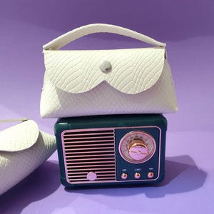 May include: Two off-white, textured, envelope-style pouches with a single top handle and a silver snap closure, positioned with one resting on top of a dark green, vintage-style radio featuring a rose gold speaker grille and circular dial.
