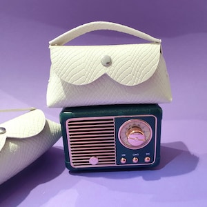 Könnte beinhalten: Eine weiße, strukturierte Clutch mit Griff und silbernem Knopfverschluss liegt auf einem teal- und roségoldfarbenen Radio im Vintage-Stil. Eine weitere ähnliche Clutch liegt an der Seite. Der Hintergrund ist einfarbig lila.