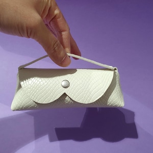 Könnte beinhalten: Ein kleines, cremefarbenes, strukturiertes Brillenetui mit geschwungenem Klappenverschluss und silbernem Knopfverschluss. Das Etui hat einen kleinen weißen Griff vor einem lila Hintergrund.