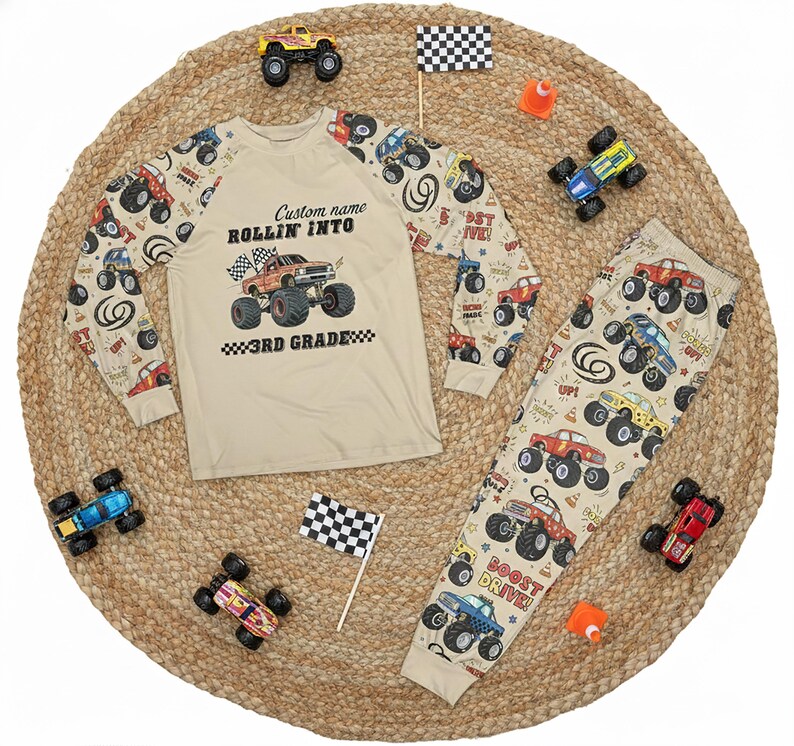 May include: A beige pajama set with a long-sleeved shirt and pants, featuring a monster truck theme. The shirt has a graphic that says "ROLLIN' INTO 3RD GRADE" and the pants have a repeating monster truck pattern. Toy monster trucks and checkered flags are scattered around.