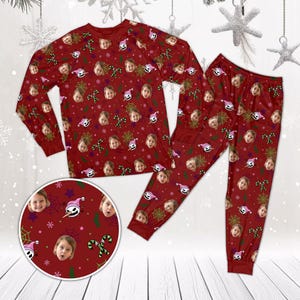 May include: A long-sleeve shirt and matching pants set in deep red, featuring a repeating pattern of a child's face, a cartoon character wearing a pink hat, green candy canes, yellow spider webs, and small white snowflakes across the fabric.
