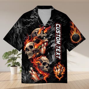 Peut inclure: Chemise de bowling noire avec un motif de crânes et de flammes. La chemise a un col en V et des manches courtes. Le motif comprend des crânes, des quilles et une boule de bowling en feu, avec le texte "CUSTOM TEXT".