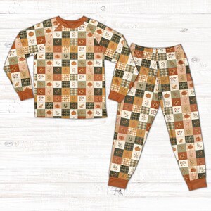 May include: A long-sleeve pajama set featuring a checkered pattern of alternating squares in shades of orange, cream, and dark green. The design includes illustrations of pumpkins, autumn leaves, and sprigs of berries across the fabric.