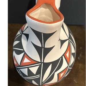 Puede incluir: Una jarra de cerámica blanca con base redondeada y cuello estrecho. La jarra está decorada con un patrón geométrico en negro y naranja. El borde de la jarra es naranja.