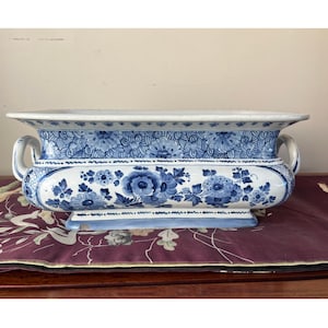 May include: A rectangular, white and blue ceramic planter with a floral design. The planter features blue floral patterns and scroll handles. The base is a light blue colour. The planter is sitting on a maroon surface.