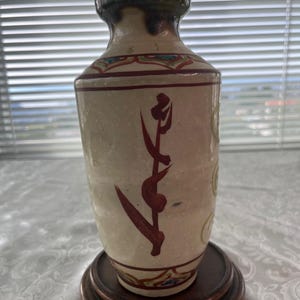 May include: A ceramic vase with a cream-colored body, adorned with maroon and green floral designs and calligraphy. The vase sits on a dark wooden stand. The rim is dark green. The vase is approximately 8 inches tall.
