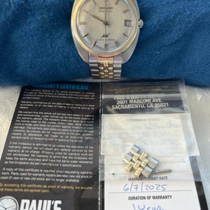 Op de afbeelding: Een vintage Longines Ultra-Chron horloge met een zilveren wijzerplaat, een goudkleurige lunette en een tweekleurige roestvrijstalen armband. De wijzerplaat toont de datum en wordt vergezeld door een garantiecertificaat en extra horlogeschakels.
