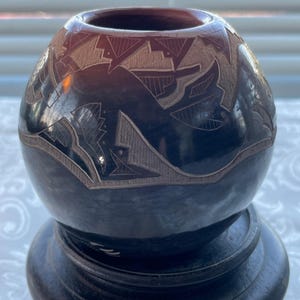 May include: A dark brown, round ceramic vase with etched geometric designs, resting on a matching wooden stand. The vase features a wide opening and a smooth, polished finish, showcasing a blend of traditional craftsmanship and artistic detail.