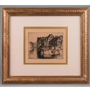 May include: A framed etching depicting a riverside scene with half-timbered houses. The artwork is encased in a textured gold frame with a cream-colored mat and a thin gold inner border. The black and white print shows buildings along a canal.