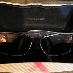 Może przedstawiać: Czarne okulary przeciwsłoneczne z prostokątną oprawką, w otwartym etui. Etui jest czarne w środku z nazwą marki "BURBERRY". Etui ma beżowo-różowy wzór w kratę na zewnątrz.