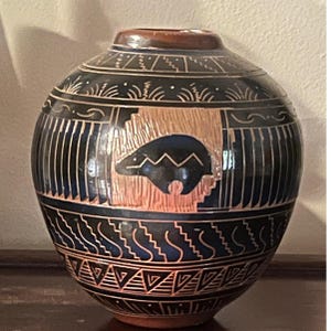 Può includere: Un vaso rotondo in ceramica fatto a mano con una combinazione di colori blu scuro e marrone. Il vaso presenta un disegno di orso e motivi geometrici. La parte superiore e il bordo sono marroni. Il vaso è probabilmente utilizzato per scopi decorativi.