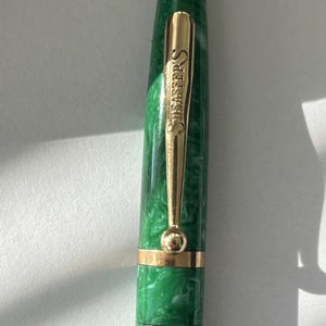 May include: A green and white marbled fountain pen with a gold-colored clip and accents. The clip is engraved with the word "SHEAFFER'S". The pen has a classic design and a shiny finish.