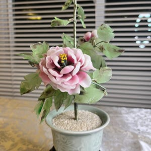 May include: A decorative artificial flower arrangement featuring a large pink peony with yellow stamens, green leaves, and a bud. The flower is in a light green pot with a decorative base, filled with small white stones.