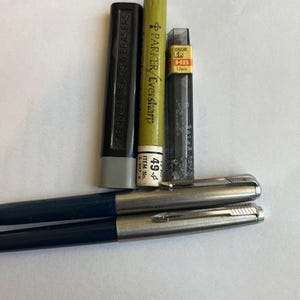 Puede incluir: Un surtido de útiles de escritura, que incluye un contenedor negro de minas y gomas de borrar Parker, un tubo de minas Parker Eversharp verde y amarillo y un contenedor negro de recarga de minas. También hay dos bolígrafos plateados y azules.