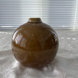 May include: A round, brown ceramic vase with a glossy finish. The vase has a small, cylindrical opening at the top. The spherical shape and warm color give it a rustic, earthy aesthetic. Ideal for home decor.