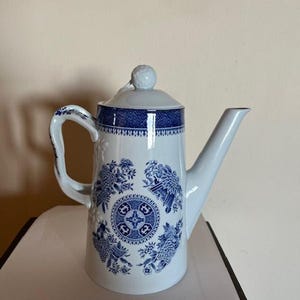 Puede incluir: Una tetera de cerámica blanca con un diseño floral azul. La tetera tiene un pico largo, un asa curva y una tapa con un pequeño pomo. El patrón floral azul se repite alrededor del cuerpo de la tetera.