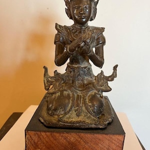 May include: A bronze-colored statue of a seated figure with ornate clothing and a tall headdress. The statue is on a black base atop a wooden block with visible wood grain. The figure's hands are clasped in a gesture of prayer or respect.