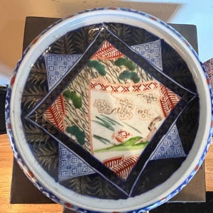 May include: A round, decorative ceramic plate with a detailed, colorful design. The plate features a central square with a landscape scene, surrounded by geometric patterns in blue, red, and green. The rim is white with a blue border.