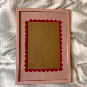 May include: A pink picture frame with a red scalloped border. The frame has a rectangular opening for a photo or artwork. The frame is made of wood and has a smooth finish. The background is a striped fabric.