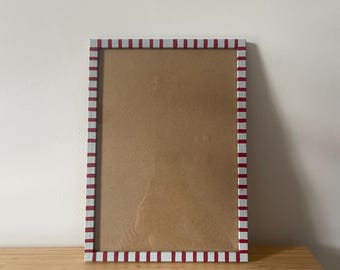 Op de afbeelding: Een rechthoekige fotolijst met een rood-witte gestreepte rand. Het frame is leeg, met een bruine achterkant zichtbaar. Het frame staat op een houten oppervlak.