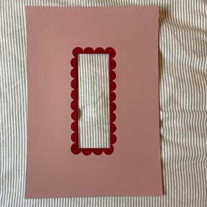 May include: A pink paper mat with a rectangular opening and a scalloped red border. The mat is on a striped fabric background. The mat is designed for displaying photos or artwork.