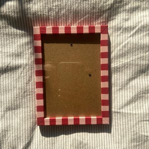 Peut inclure: Cadre photo rectangulaire avec une bordure à rayures rouges et blanches. Le cadre a un dos marron et deux petits trous pour l'accrocher. Le cadre est vide, prêt pour une photo.