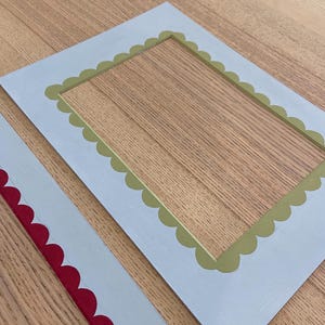 May include: Two decorative picture frames. One frame is light blue with a scalloped olive green inner border. The second frame is light blue with a scalloped red inner border. Both frames are rectangular.